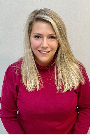 The image shows a woman posing for a professional portrait, wearing a red top and standing against a light-colored background.