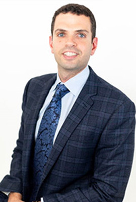 The image shows a man standing in front of a plain background. He is wearing a suit, tie, and patterned jacket.