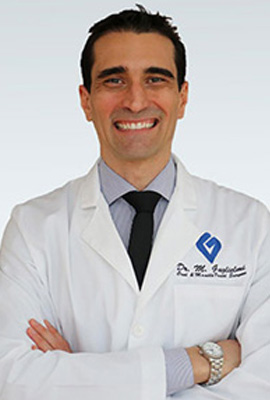 The image shows a man in a white lab coat standing against a plain background, smiling slightly and looking directly at the camera.