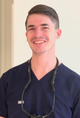 A smiling male nurse wearing a white coat and stethoscope, standing in front of a light-colored wall.