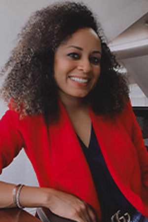 The image features a woman with curly hair, smiling at the camera. She is wearing a red blazer and is seated in front of a desk.