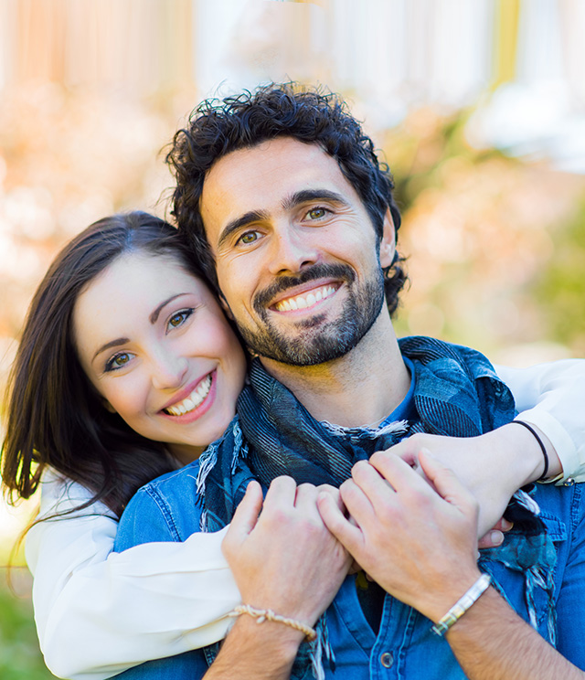 The image shows a man and woman embracing each other with smiles, set against an outdoor background.