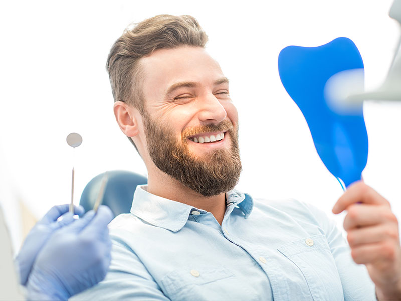 The image shows a man with a beard, holding a toothbrush and smiling broadly while sitting in a dental chair. He is wearing a blue surgical mask and has a dental mirror in his hand.
