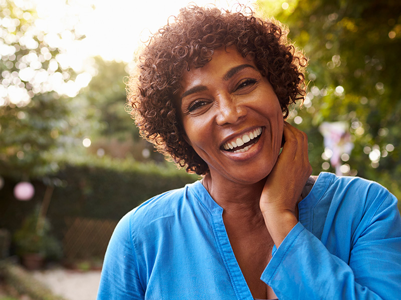 A smiling woman with short curly hair, wearing a blue sleeveless top and standing outdoors in bright sunlight.