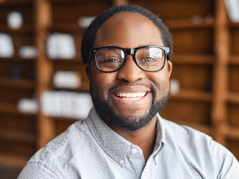 A smiling man wearing glasses, a beard, and a blue shirt, posing in front of bookshelves.