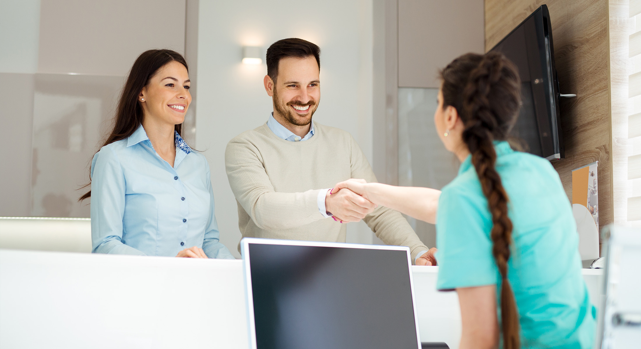 A man and a woman shaking hands in an office setting, with another person observing the interaction.