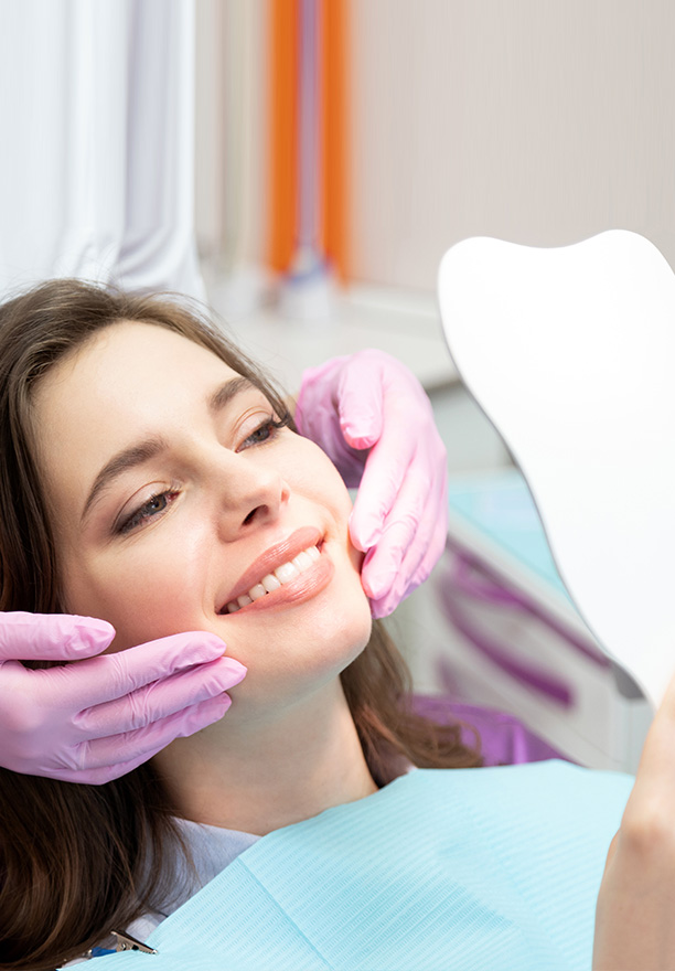 The image depicts a young woman sitting in a dental chair, receiving dental care. She is smiling and appears to be relaxed as she looks directly at the camera. A dental professional is performing a procedure on her teeth, using a dental mirror and a small handheld device. The setting suggests a modern dental office with clean and sterile equipment.