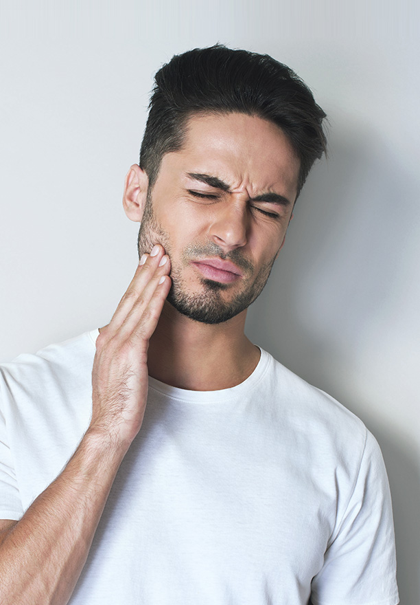Man in white shirt with hand on neck, looking directly at camera, standing against grey background.