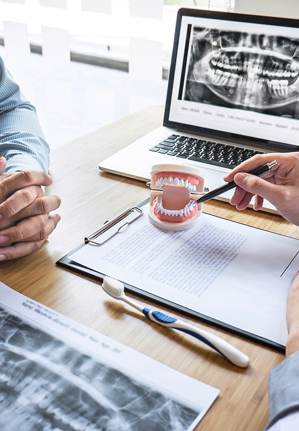 The image shows a dental professional examining a patient's teeth using a magnifying glass, with the patient seated and holding a toothbrush. They are in an office setting, possibly during a consultation or examination appointment.