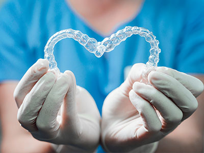 A person s hand holding a transparent heart-shaped object, likely a dental appliance, against a blue background with a medical theme.