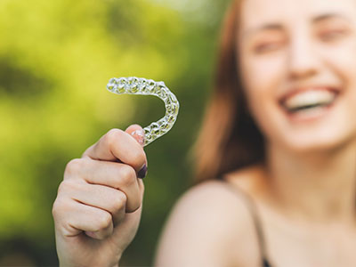 An image shows a smiling young woman holding up a clear plastic dental aligner against her teeth, with a blurred background of outdoor greenery.