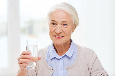 The image shows an elderly woman holding a glass of water, smiling at the camera with her thumb up.