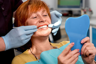 A woman is seated in a dental chair, smiling at the camera while holding a blue dental impression tray and wearing a surgical mask.