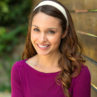 The image is a portrait of a young woman with long brown hair, wearing a purple top and a white headband. She is smiling and posing against a wooden fence in an outdoor setting.