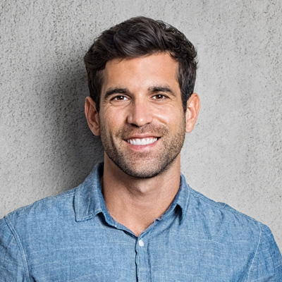 This is a color photograph of a man with short hair, smiling at the camera. He has a beard and mustache, and is wearing a light blue shirt. The background is a neutral gray tone, providing a clear contrast to the subject.