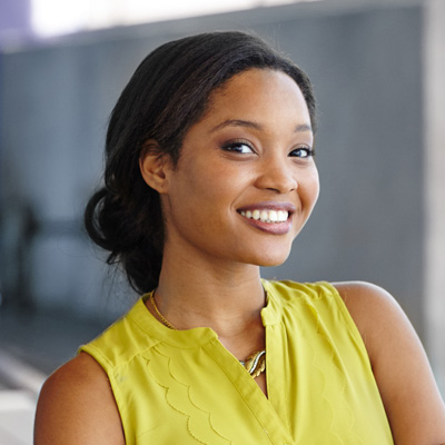 A woman with a radiant smile, wearing a yellow top and standing in front of a building.