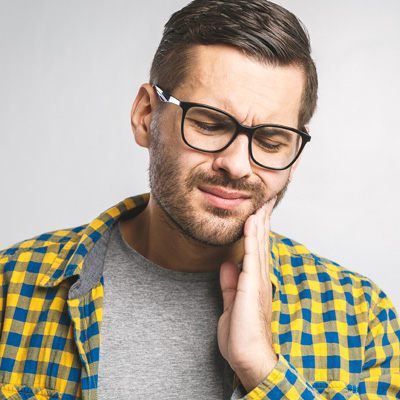 The image shows a man with a beard, wearing glasses and a plaid shirt, holding his hand to his mouth as if he s in pain or discomfort.