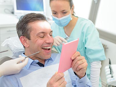 The image shows a man sitting in a dental chair, holding up a pink card with a smile on his face, while a dentist and dental hygienist are attending to him.