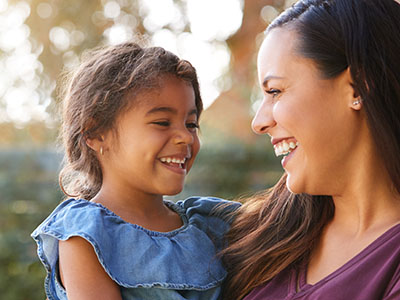 An adult woman and a young child, both smiling, share a heartfelt moment outdoors.