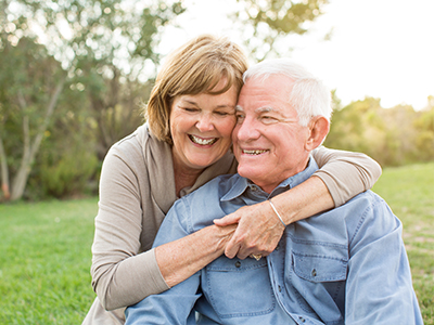 An elderly couple sharing a warm embrace in an outdoor setting, with the woman wearing a white top and the man in a blue shirt.