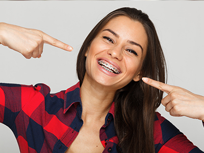 The image shows a woman with a wide smile, pointing to her teeth with one hand and holding up a finger with the other. She has long hair, is wearing a red plaid shirt, and has a playful expression on her face.