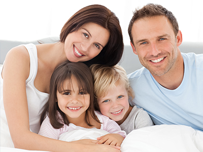 The image shows a family of four, including two adults and two children, posing for a photo in a bedroom setting with a cozy atmosphere.