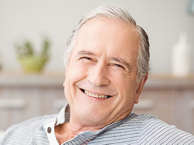 The image shows a smiling older man with gray hair, wearing glasses and a white shirt, sitting in a relaxed pose with his hands resting on his chest.